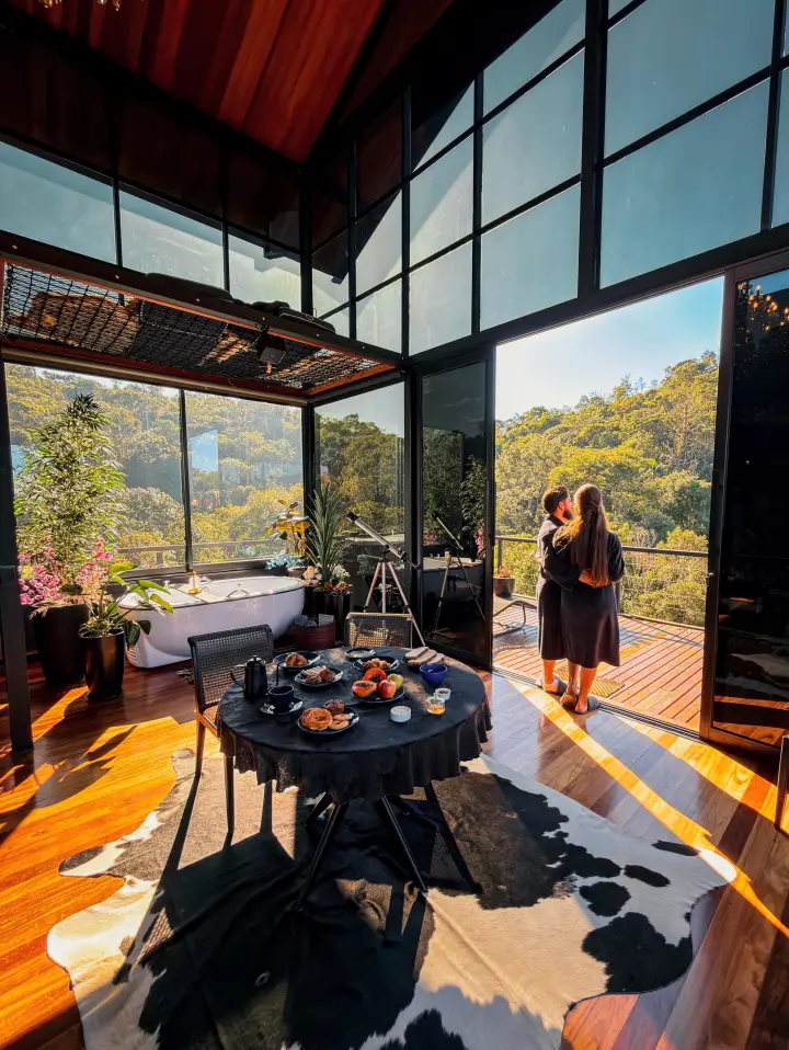 Casal abraçado na varanda de um chalé envidraçado, à mesa do café da manhã