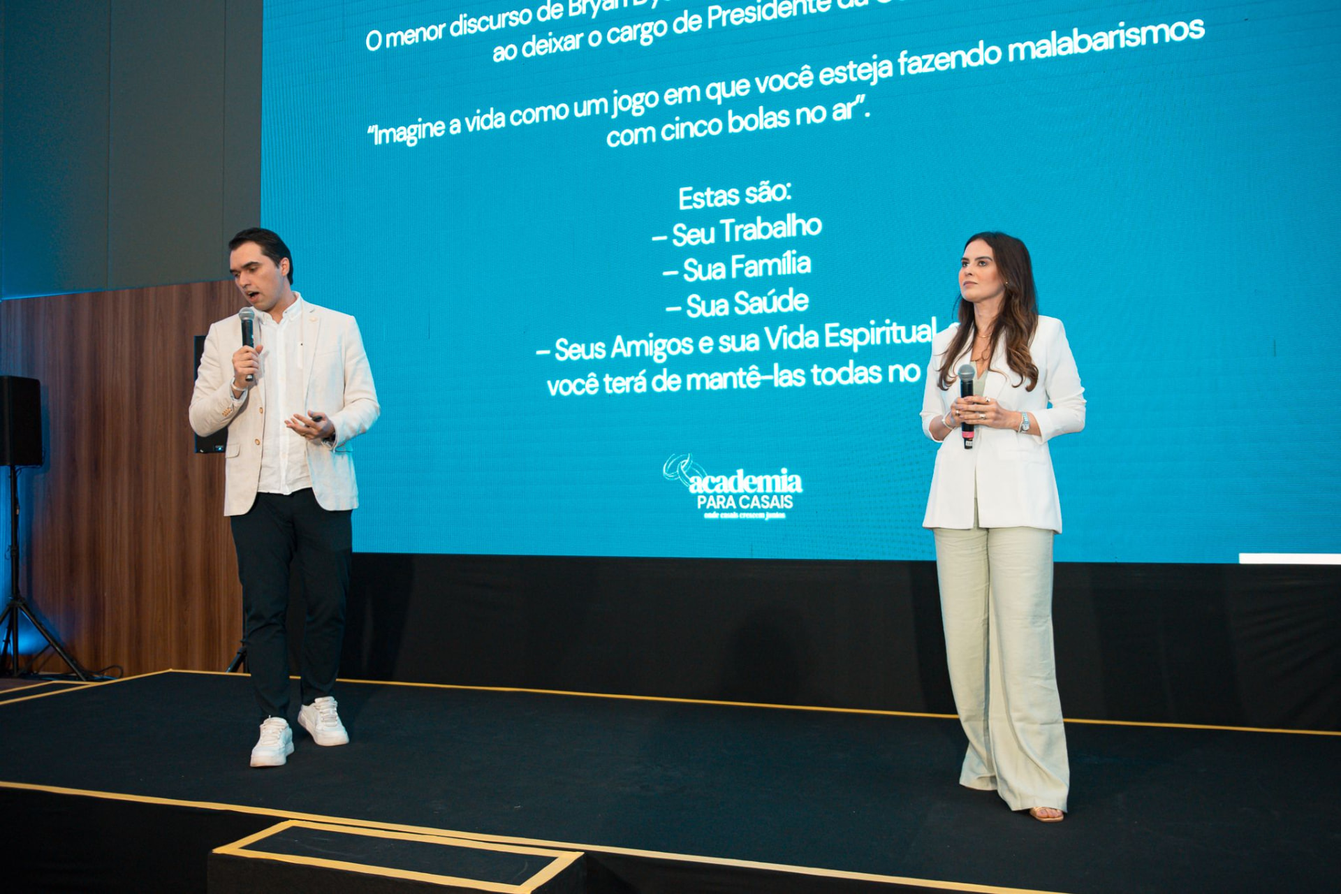 Marcos e Viviane conduzindo um encontro com microfones diante de um telão, com os casais sentados às mesas